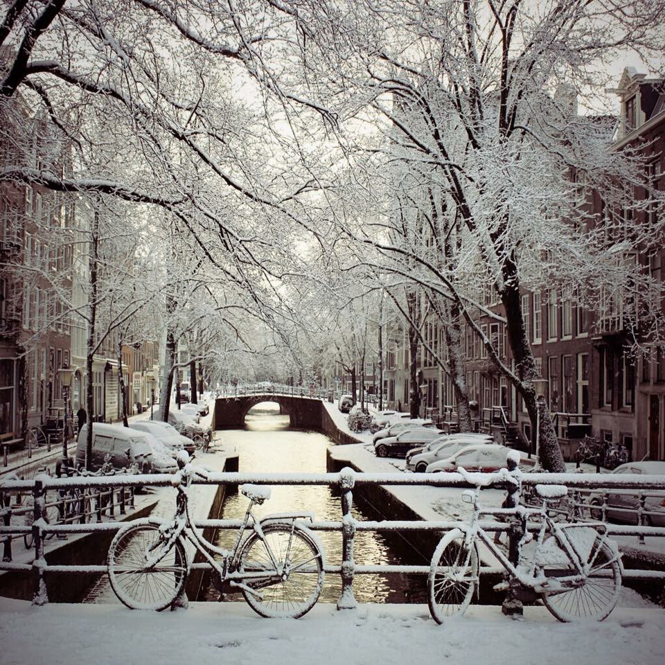 Snow-covered bicycles on a bridge over a frozen Amsterdam canal at dusk